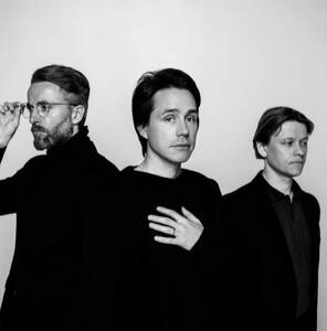A black and white photo of Mew from 2017. The three members stand together, dressed in black.